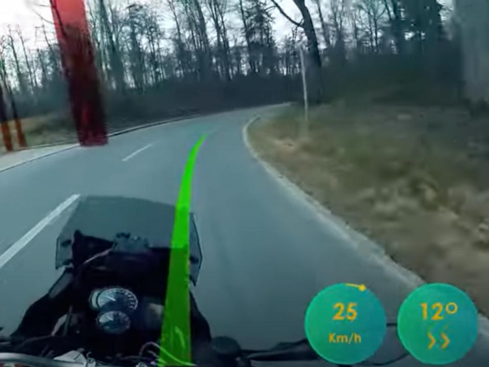 <p>Aegis Rider HUD käyttää modernia sähköistä paikanninteknologiaa, kameroita ja keinoälyä (AI, Artificial Intelligence) esittääkseen optimaalisen ajolinjan ja -nopeuden kuljettajan näkökenttään. Punainen varoituspylväs kertoo jalankulkijasta.</p>